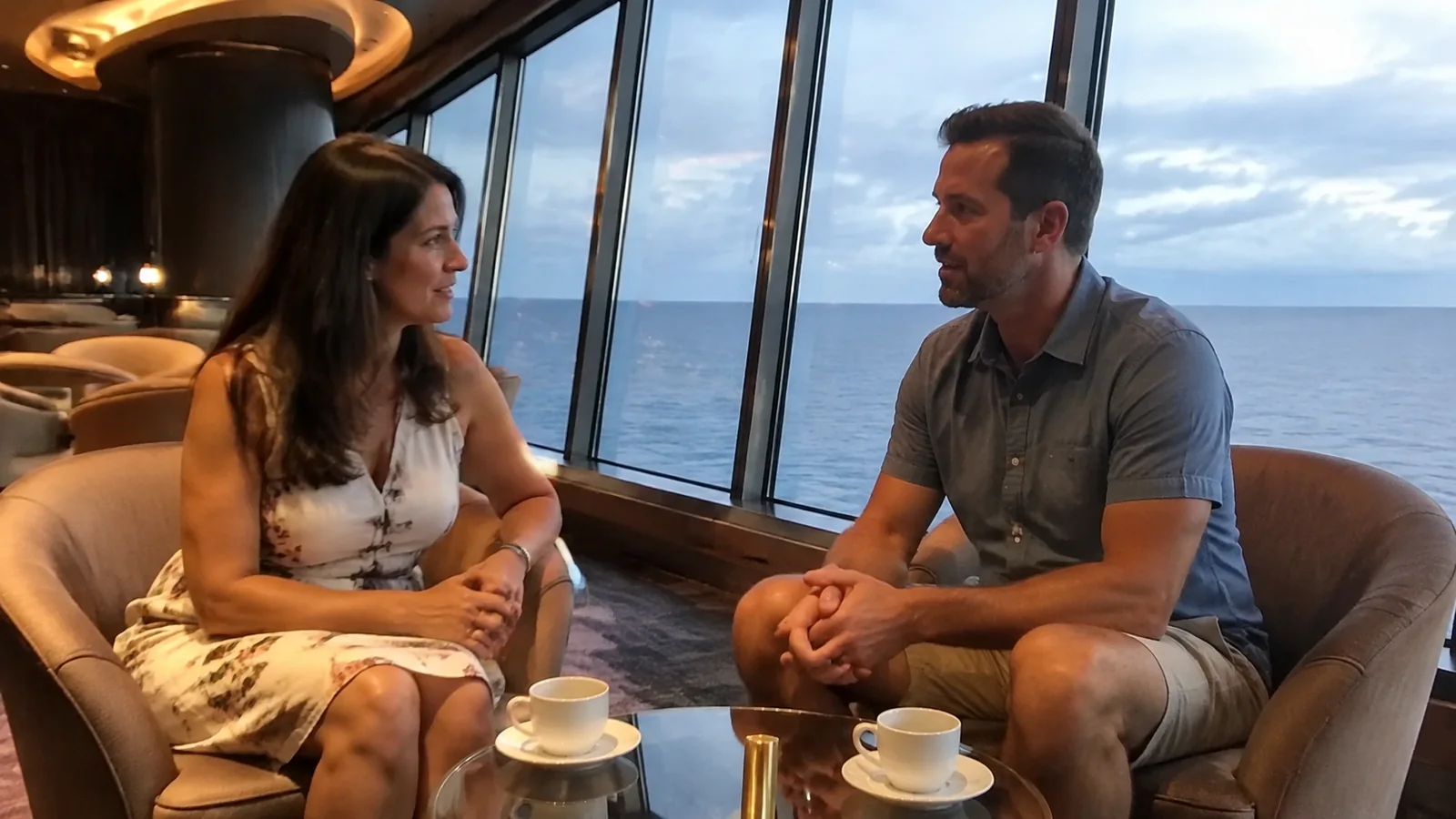 Couple talks in a lounge beside slanted ocean windows.