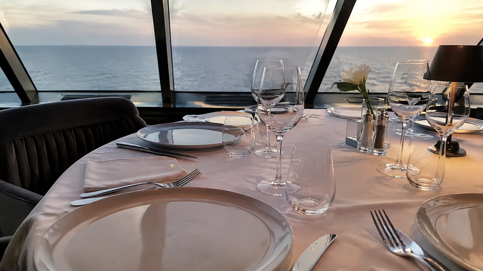 White plates and glassware are arranged on a dining table by the sea.