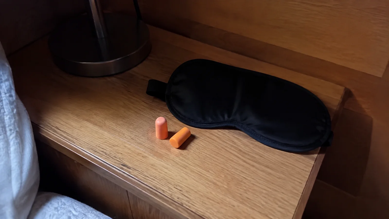Black sleep mask and orange earplugs on a wooden bedside table.