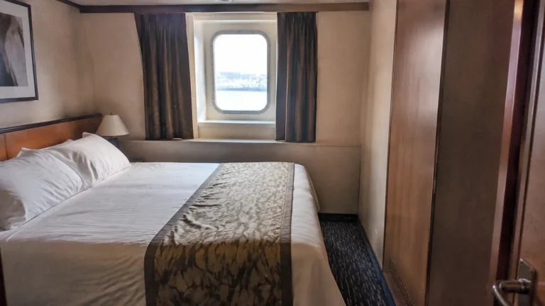 Cruise cabin with a double bed, curtained window, and wooden wardrobe