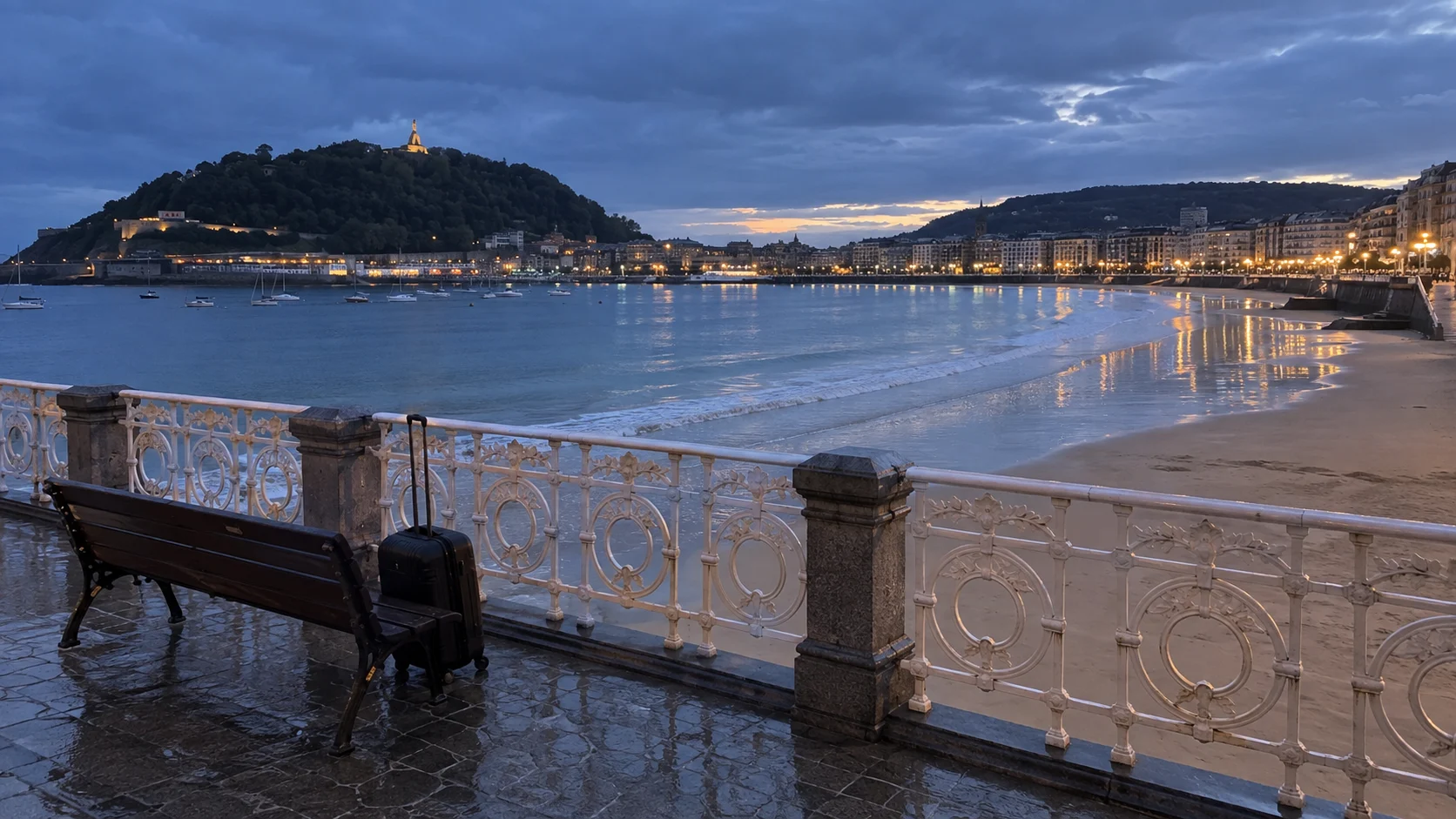 Rolling suitcase beside the rain-wet promenade overlooking La Concha Bay in San Sebastián at blue hour.