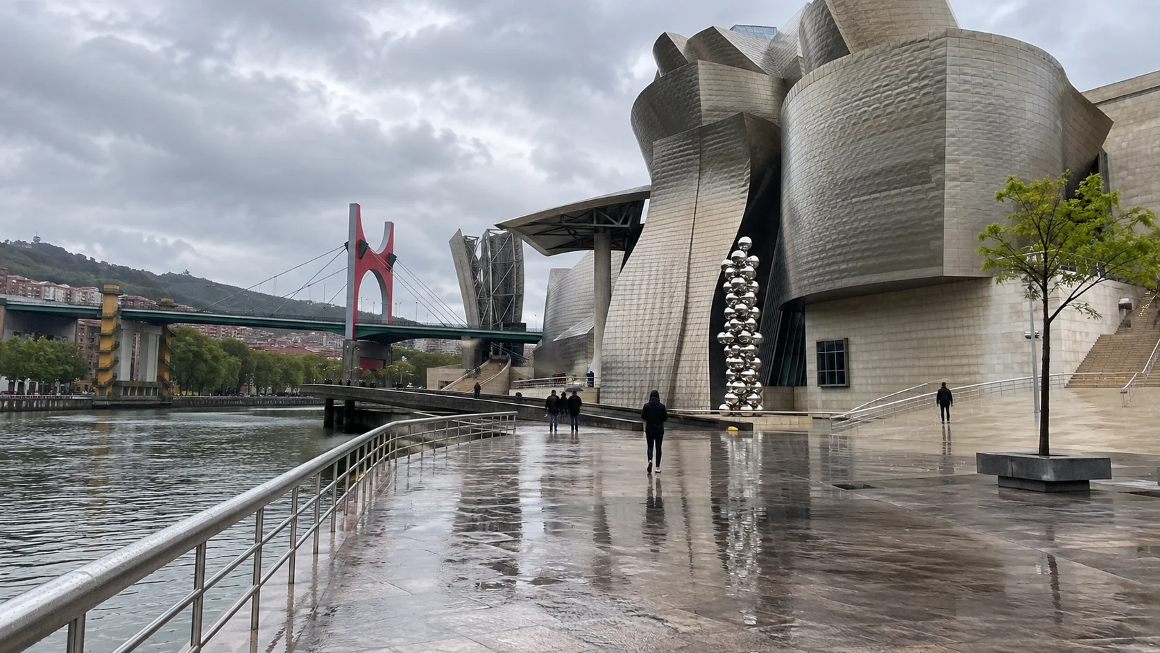 Guggenheim Bilbao and the red La Salve Bridge reflected on rain-wet riverside pavement.