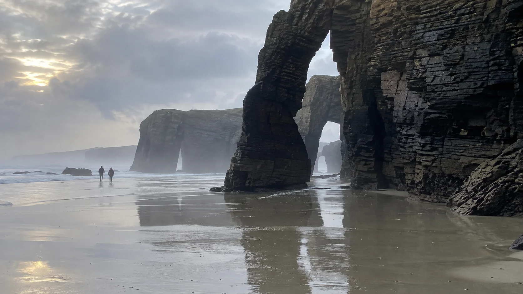 Walkers beneath the towering sea arches of Playa de las Catedrales at low tide.
