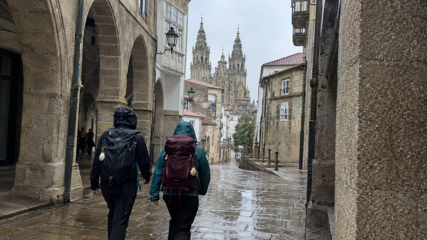 Two rain-clad pilgrims walking toward Santiago Cathedral through the historic stone center.
