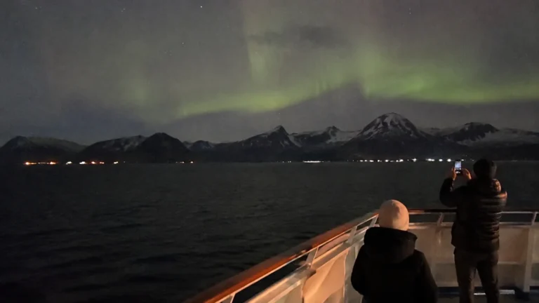 Passengers photographing northern lights above snowy mountains from a ship's deck
