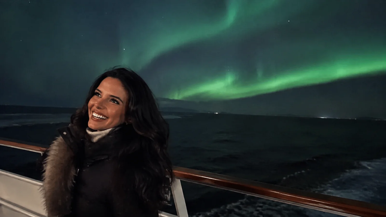 Smiling woman on a ship's deck beneath green northern lights