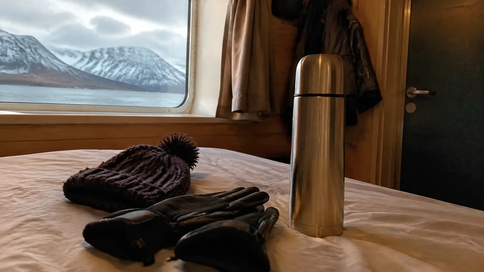 Thermos, knit hat, and gloves on a cruise cabin bed beside a mountain-view window