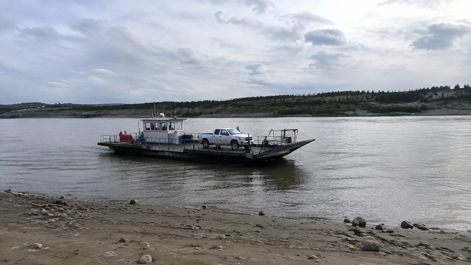 Small vehicle ferry crossing calm water near a rocky bank.