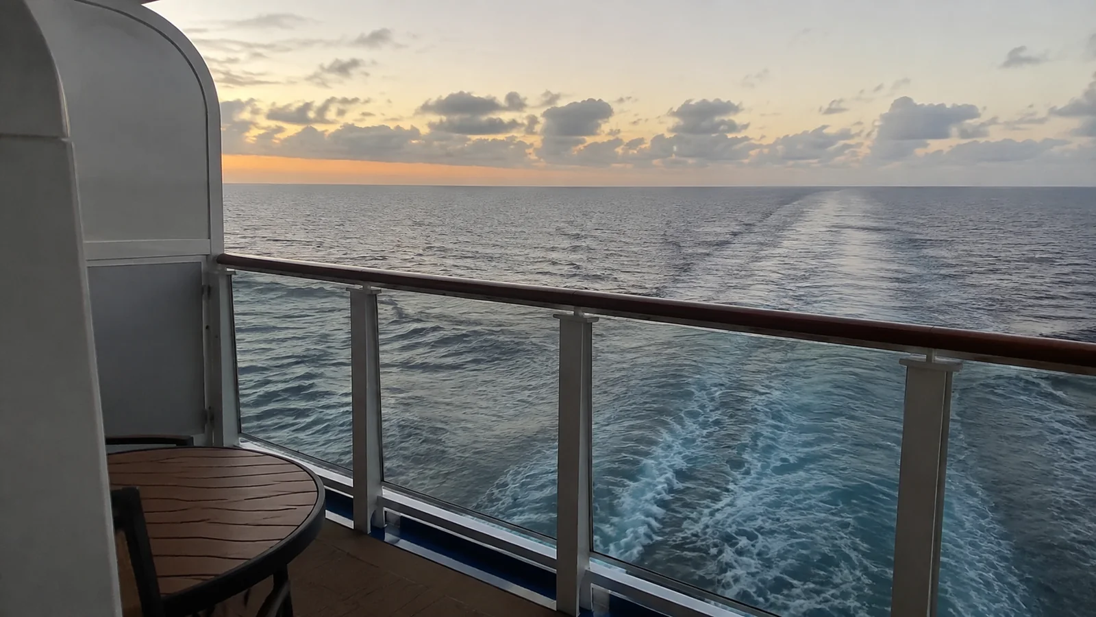 Ship's wake stretches toward the sunset beyond a balcony railing.