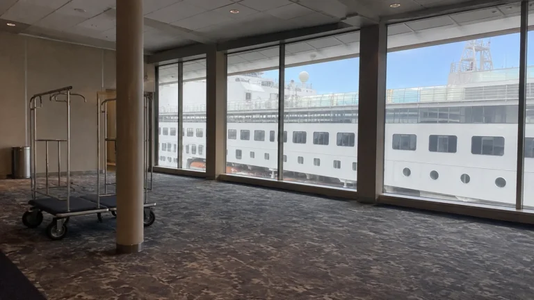Luggage cart stands in a terminal beside windows facing a cruise ship.
