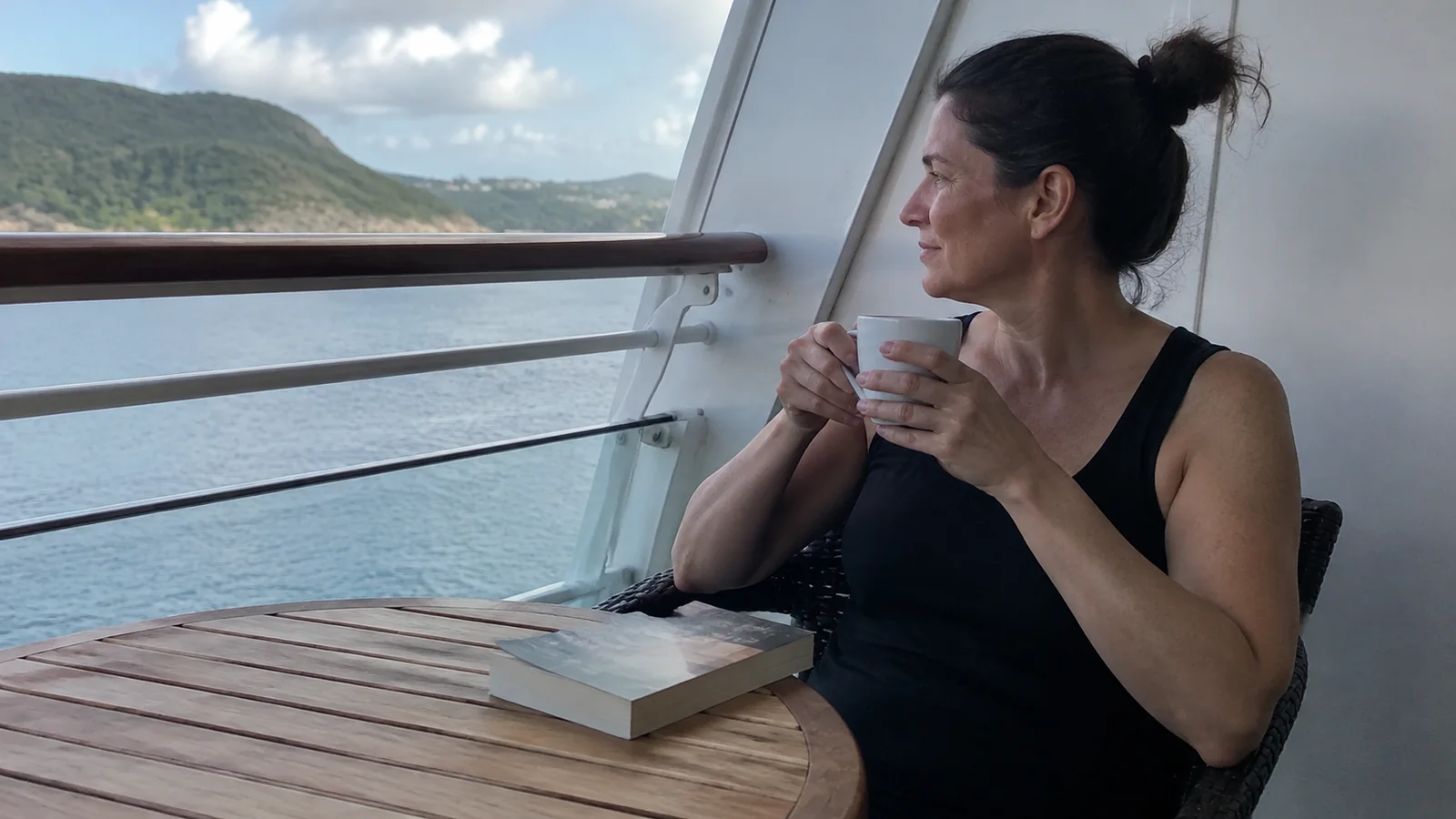 Woman holds a cup on a balcony overlooking a green coastline.