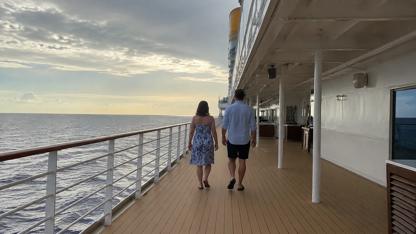Couple walks along an outdoor ship promenade at sunset.