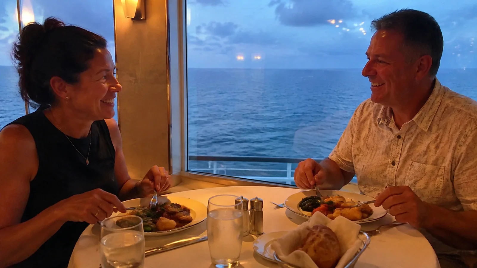 Couple dines beside a window overlooking the ocean at dusk.