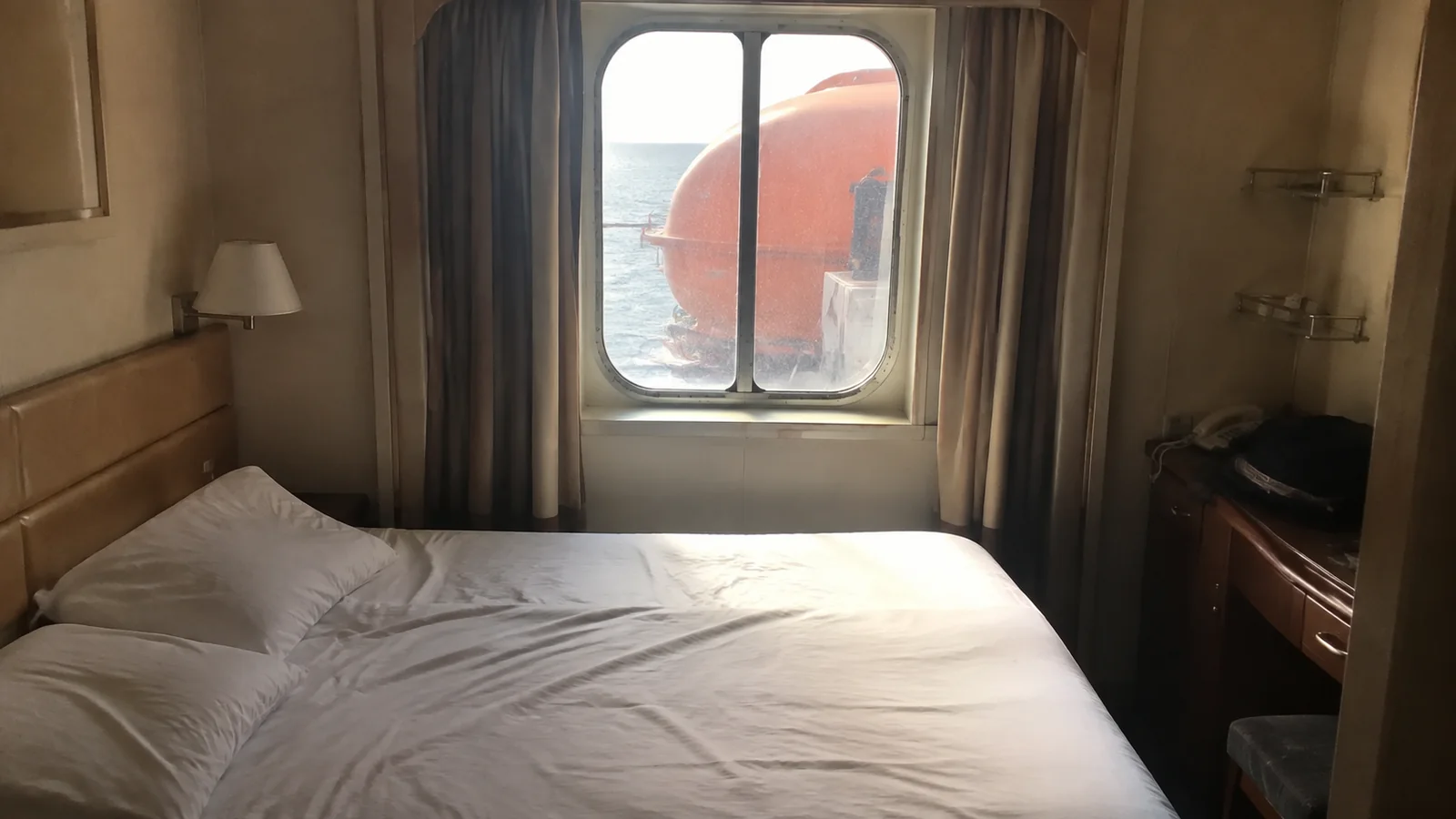 Cabin bed beneath a window partly blocked by an orange lifeboat.