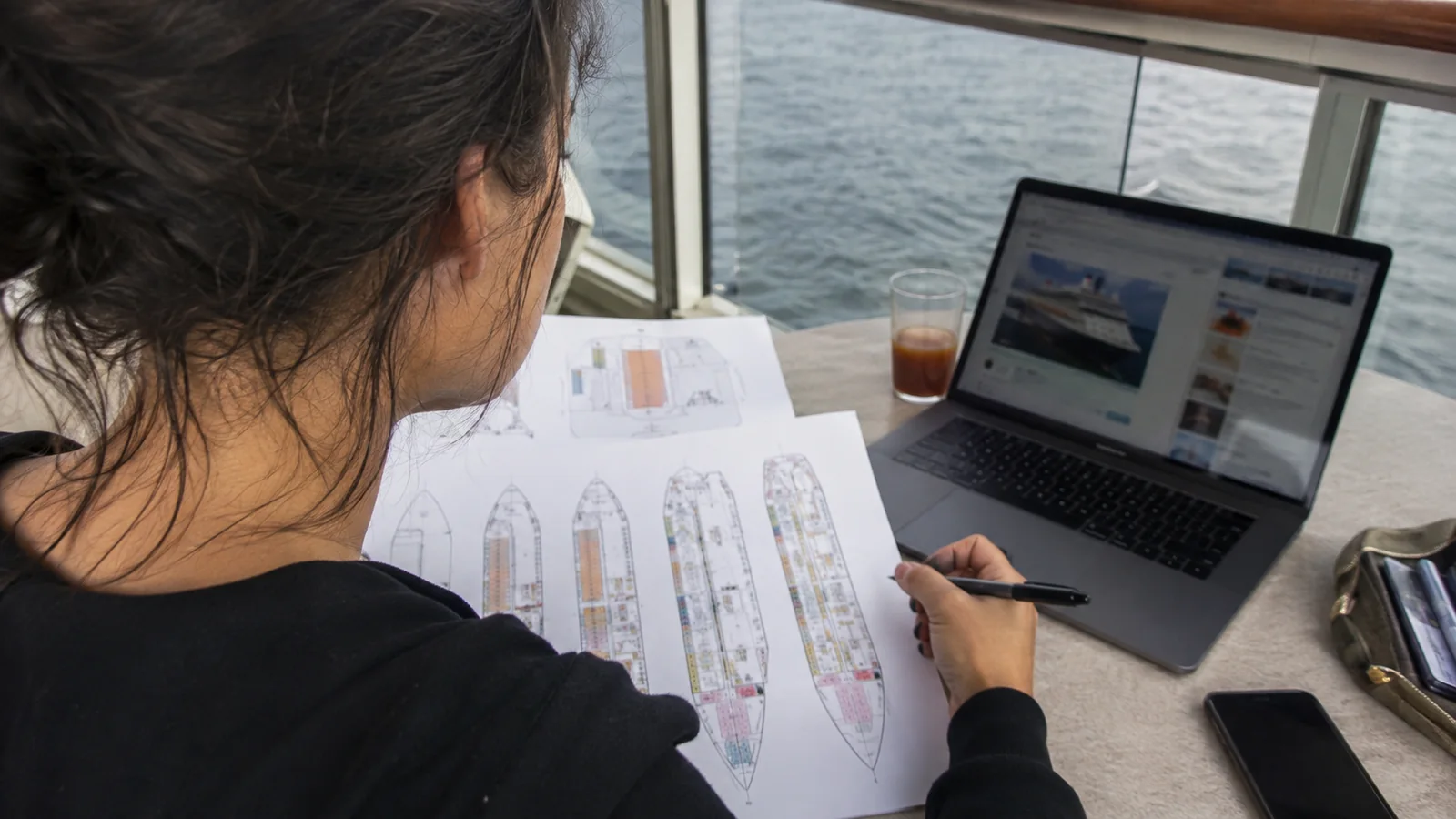 Woman reviewing printed cruise ship deck plans beside a laptop at a window