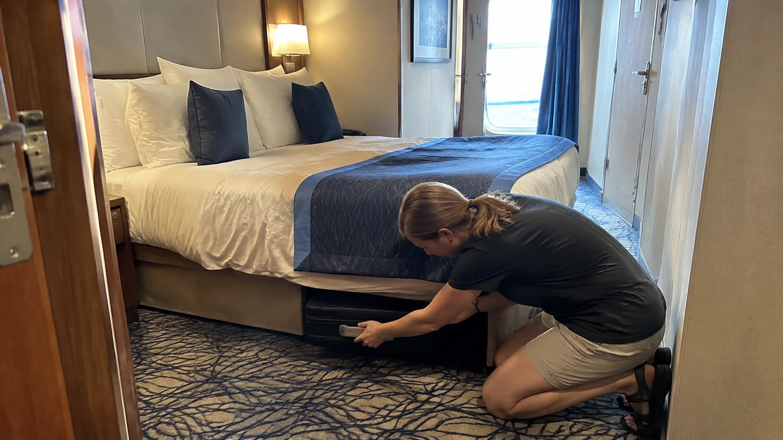 Traveler crouches to slide a suitcase beneath a stateroom bed.