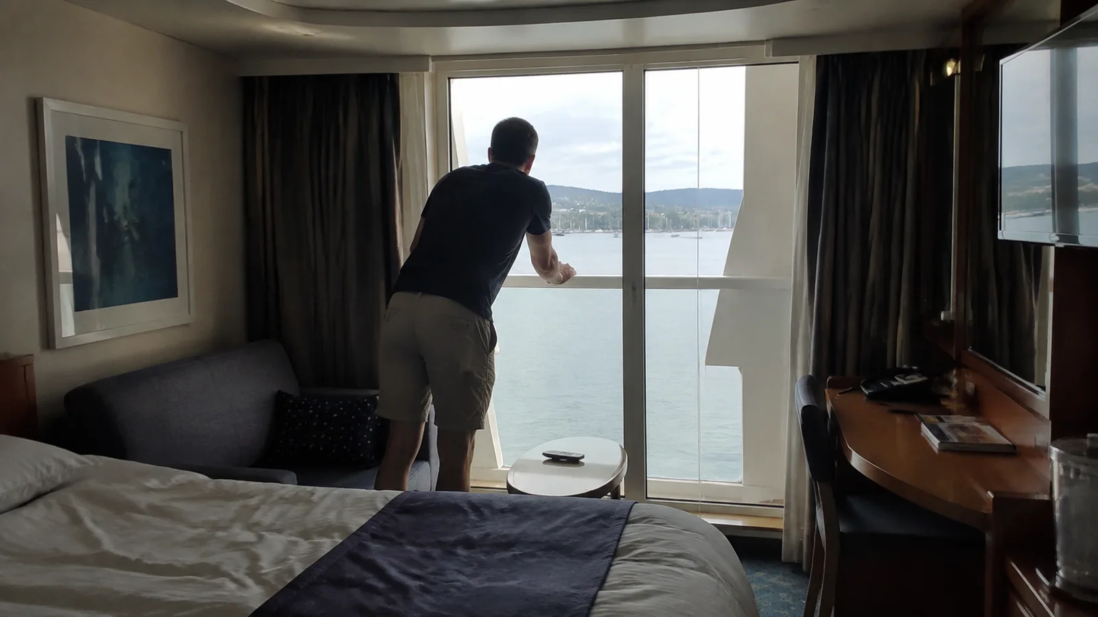 Man looking through a cruise cabin balcony door toward the harbor