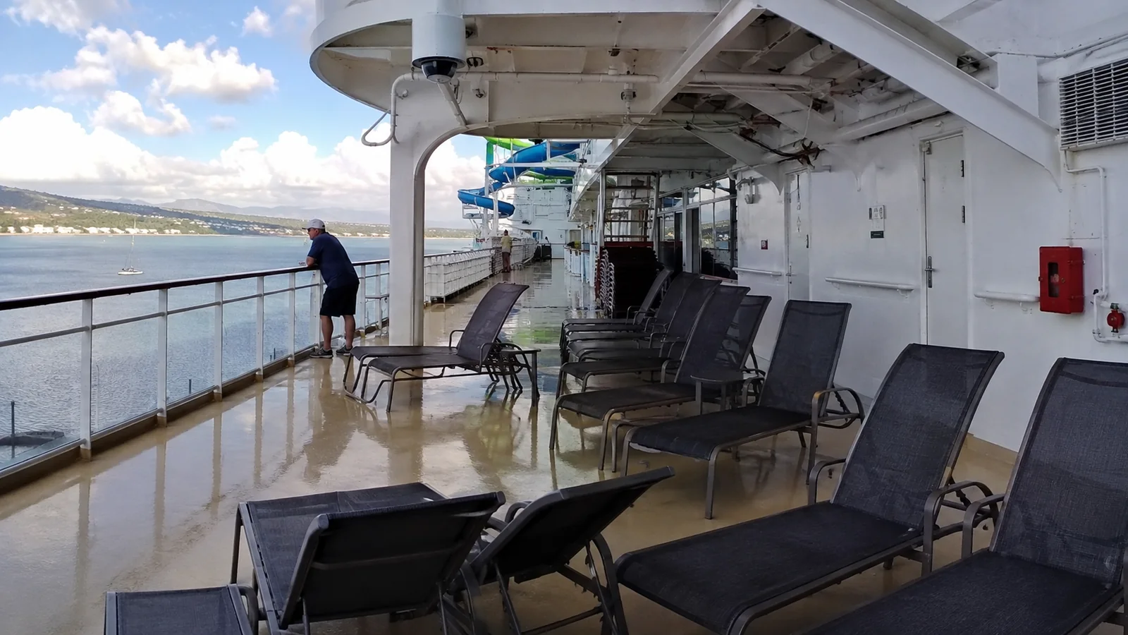 Lounge chairs on a wet cruise ship promenade with a passenger at the railing