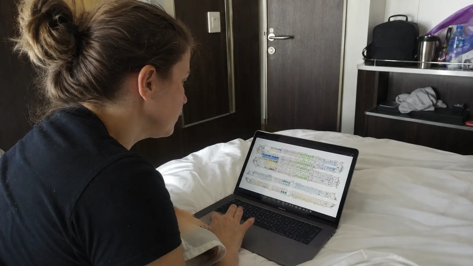 Woman using a laptop on a cruise cabin bed with a deck plan on screen