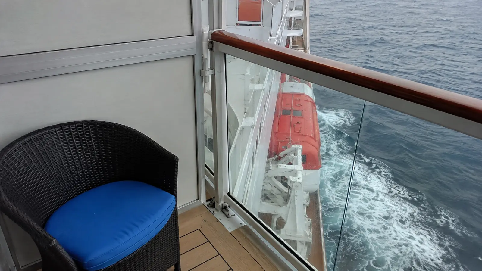Blue chair on a cruise ship balcony with a lifeboat and ship wake below
