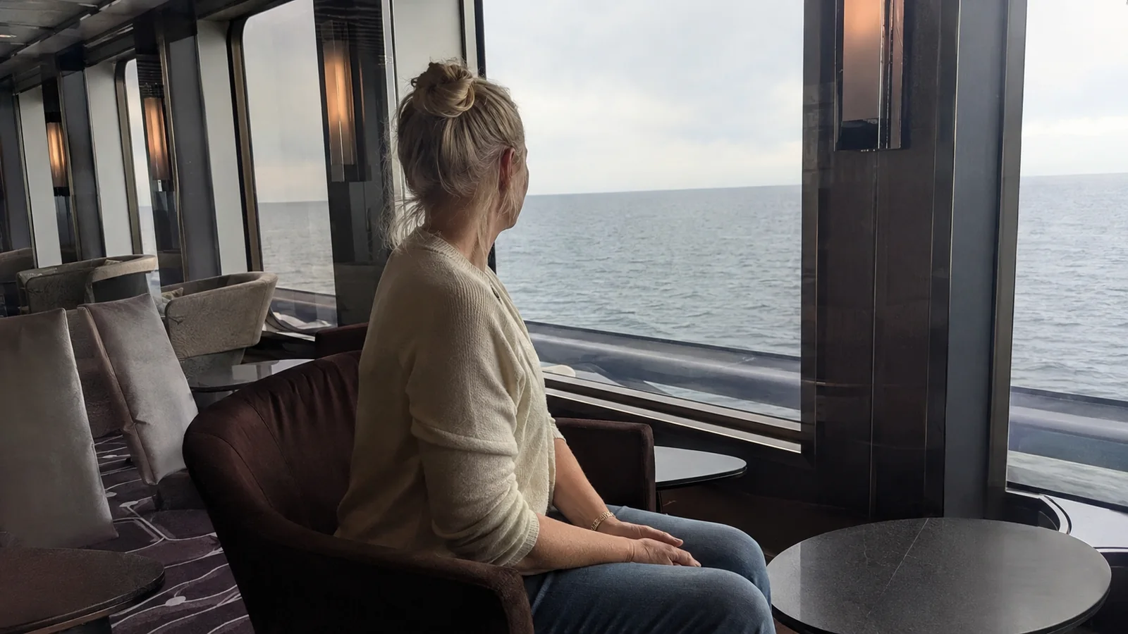 Woman sits beside a large ship window overlooking the sea.