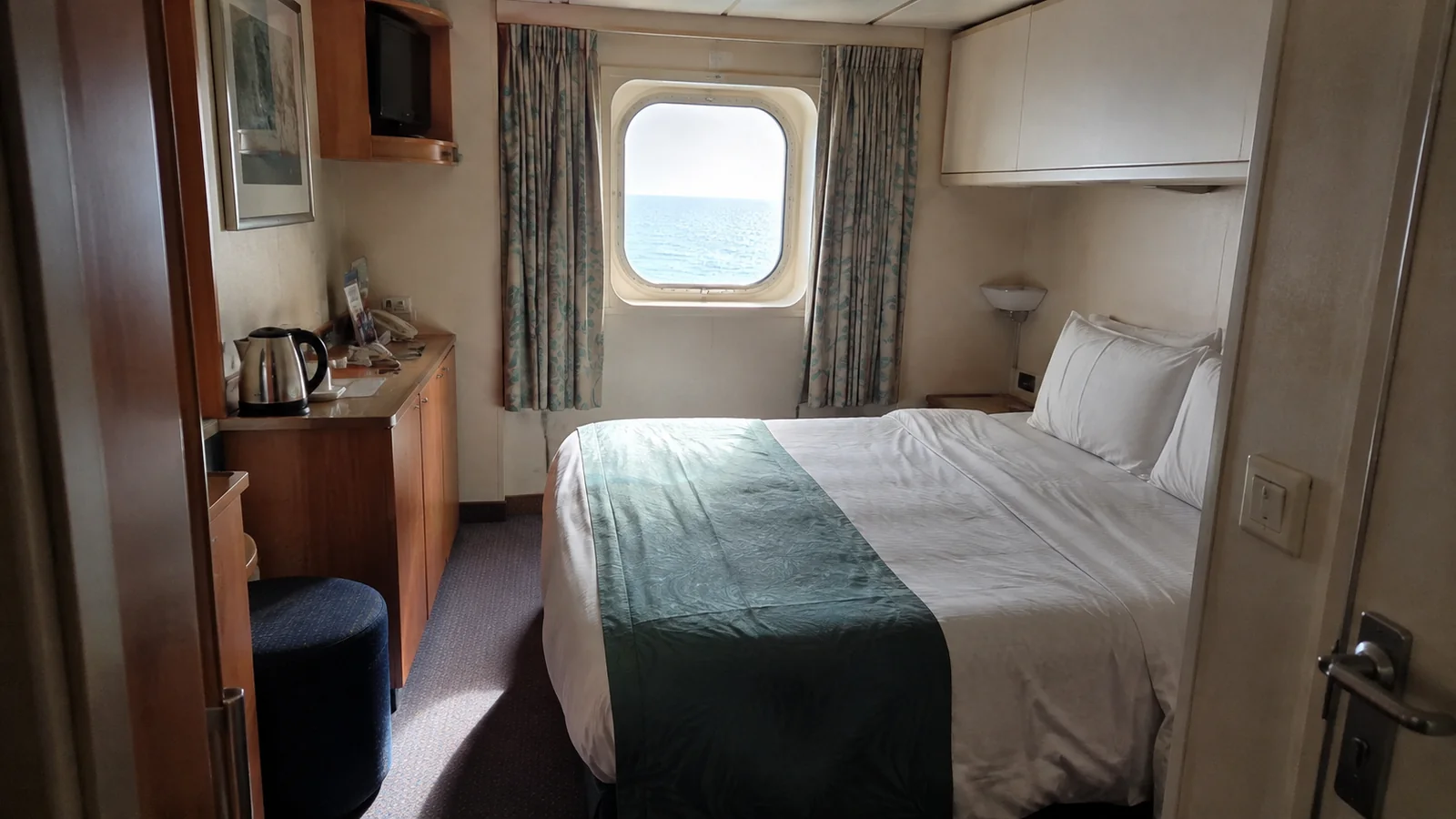 Compact cruise cabin with a double bed, desk, and ocean-view window