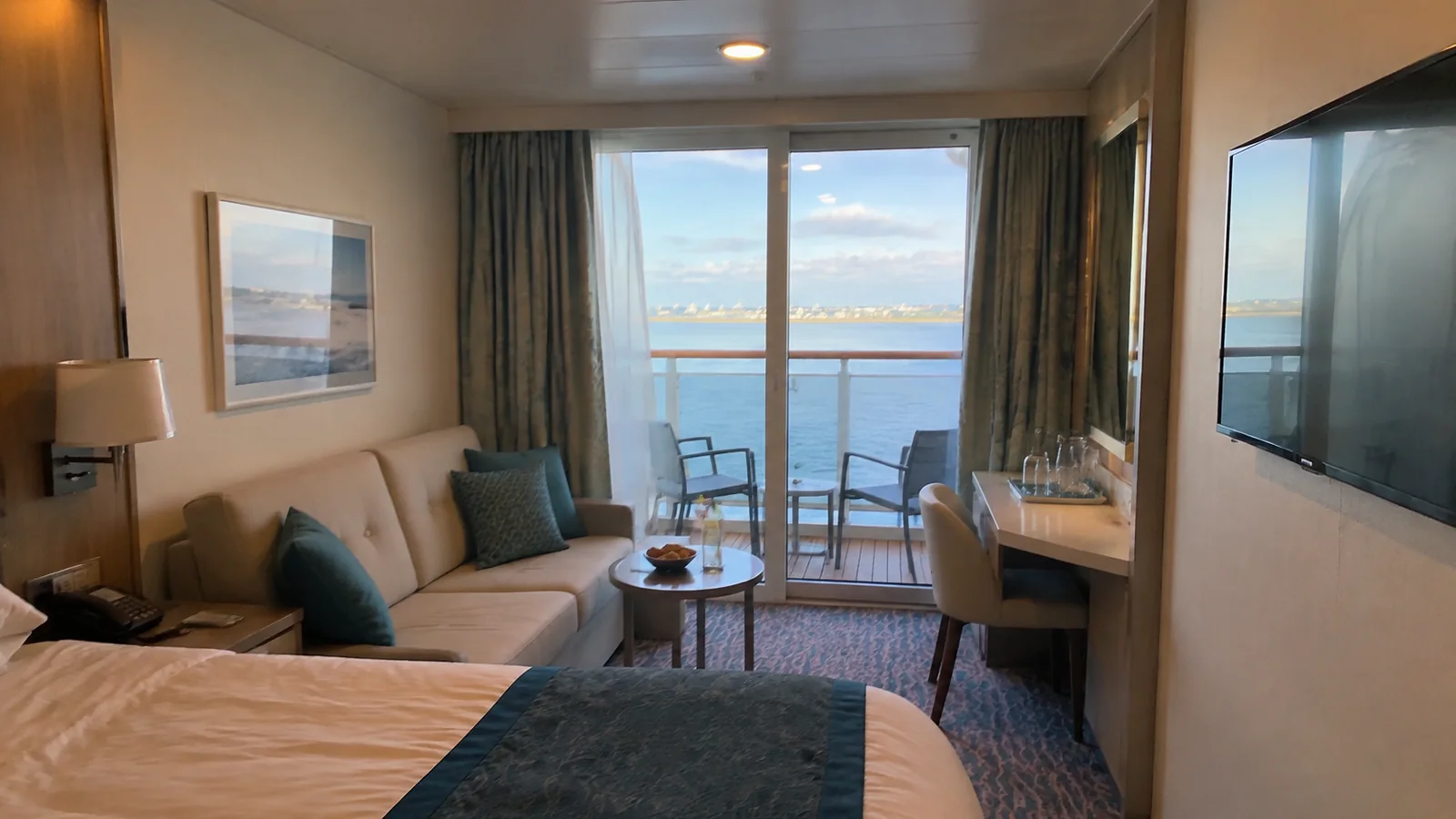 Stateroom sofa and desk face glass balcony doors at sunset.