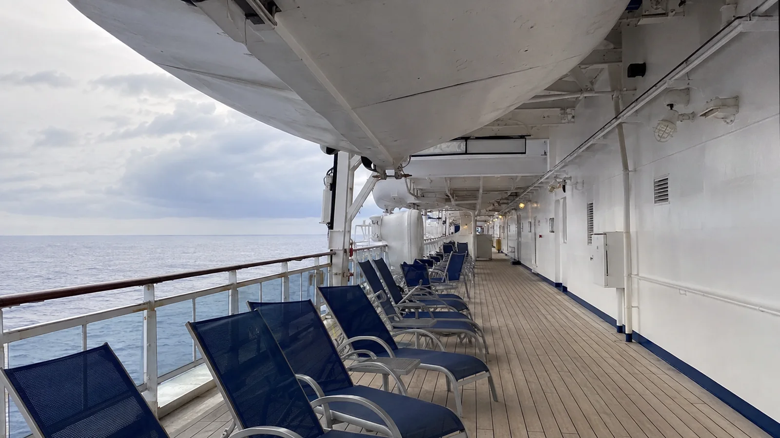 Blue lounge chairs lining a cruise ship promenade beneath lifeboats