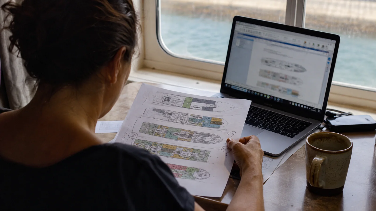 Woman comparing printed cruise ship deck plans with a laptop beside a window