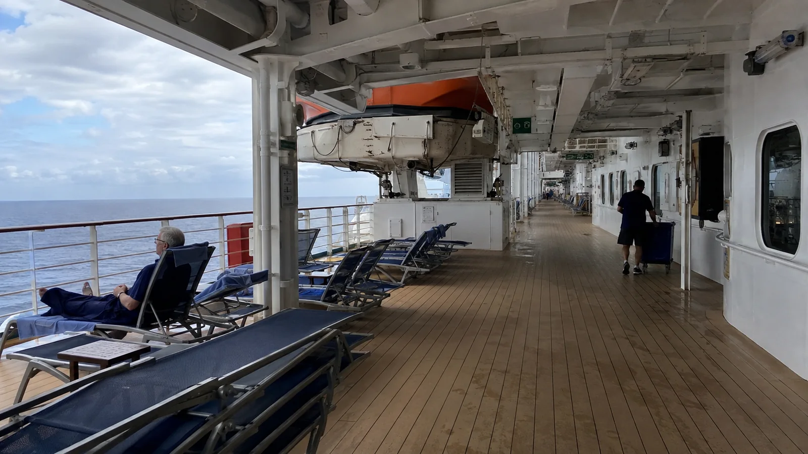 Passengers on a wooden cruise ship promenade beneath lifeboats