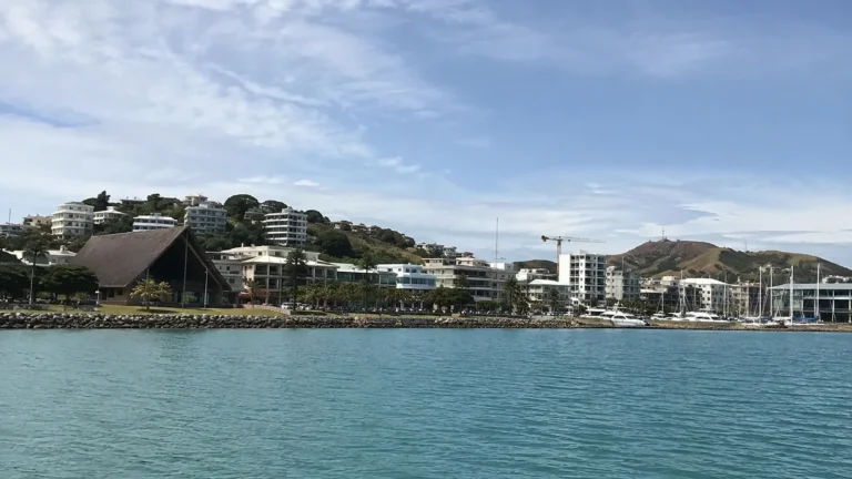 Noumea waterfront with marina masts and low buildings beneath green hills.