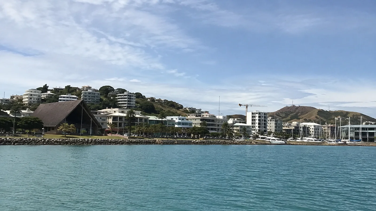 Noumea waterfront with marina masts and low buildings beneath green hills.