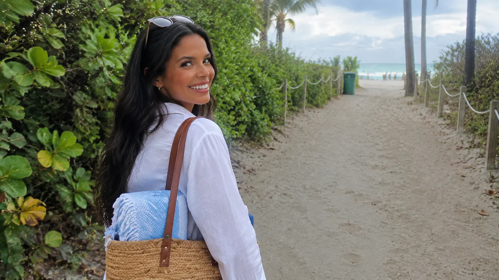 Smiling woman carrying a towel and beach bag along a sandy beach path