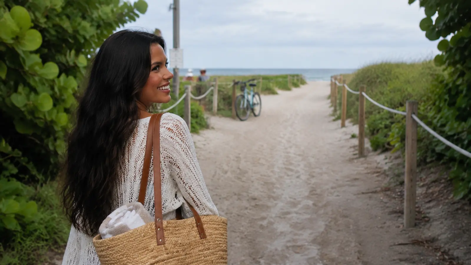 Smiling woman carrying a beach bag along a sandy path toward the shore