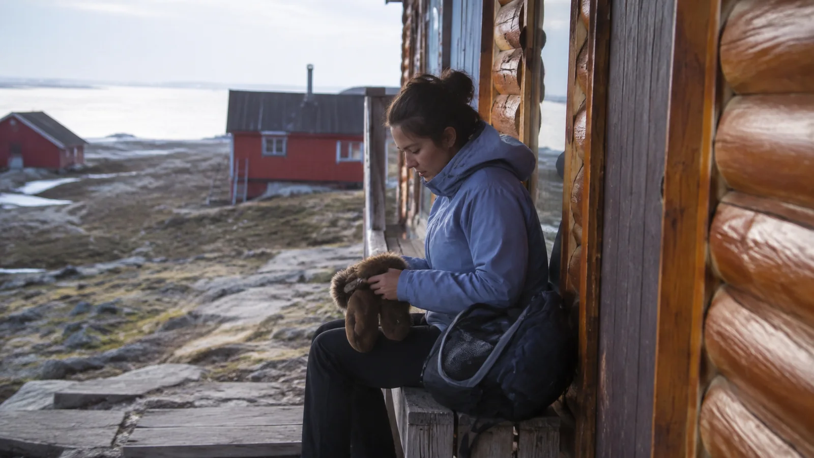 Woman checking boots outside a wooden cabin in a rocky settlement.
