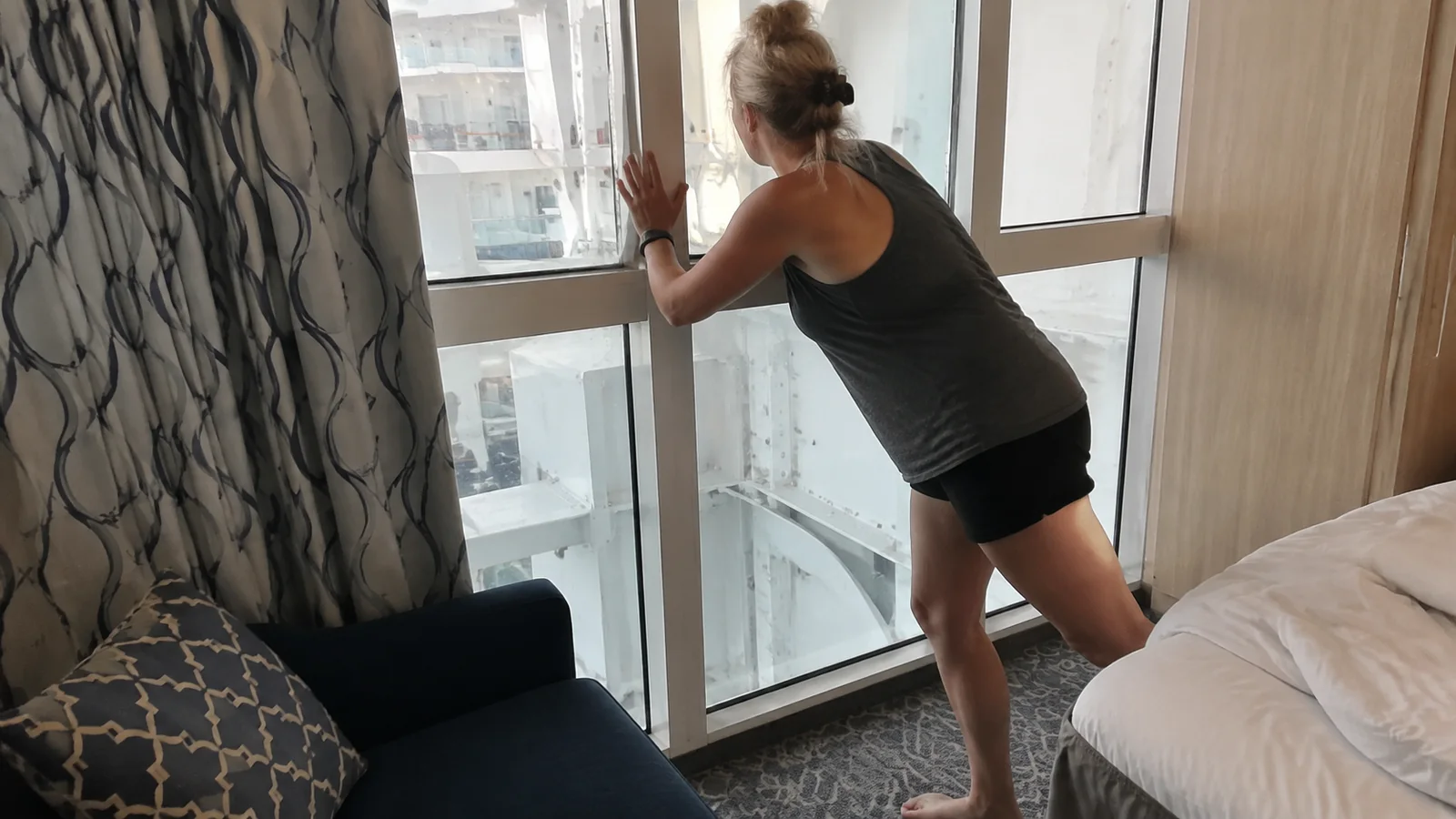 Woman looking through a cruise cabin balcony door toward an exterior obstruction