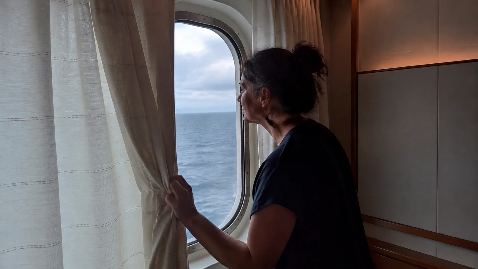 Woman holds a curtain aside beside a stateroom window.