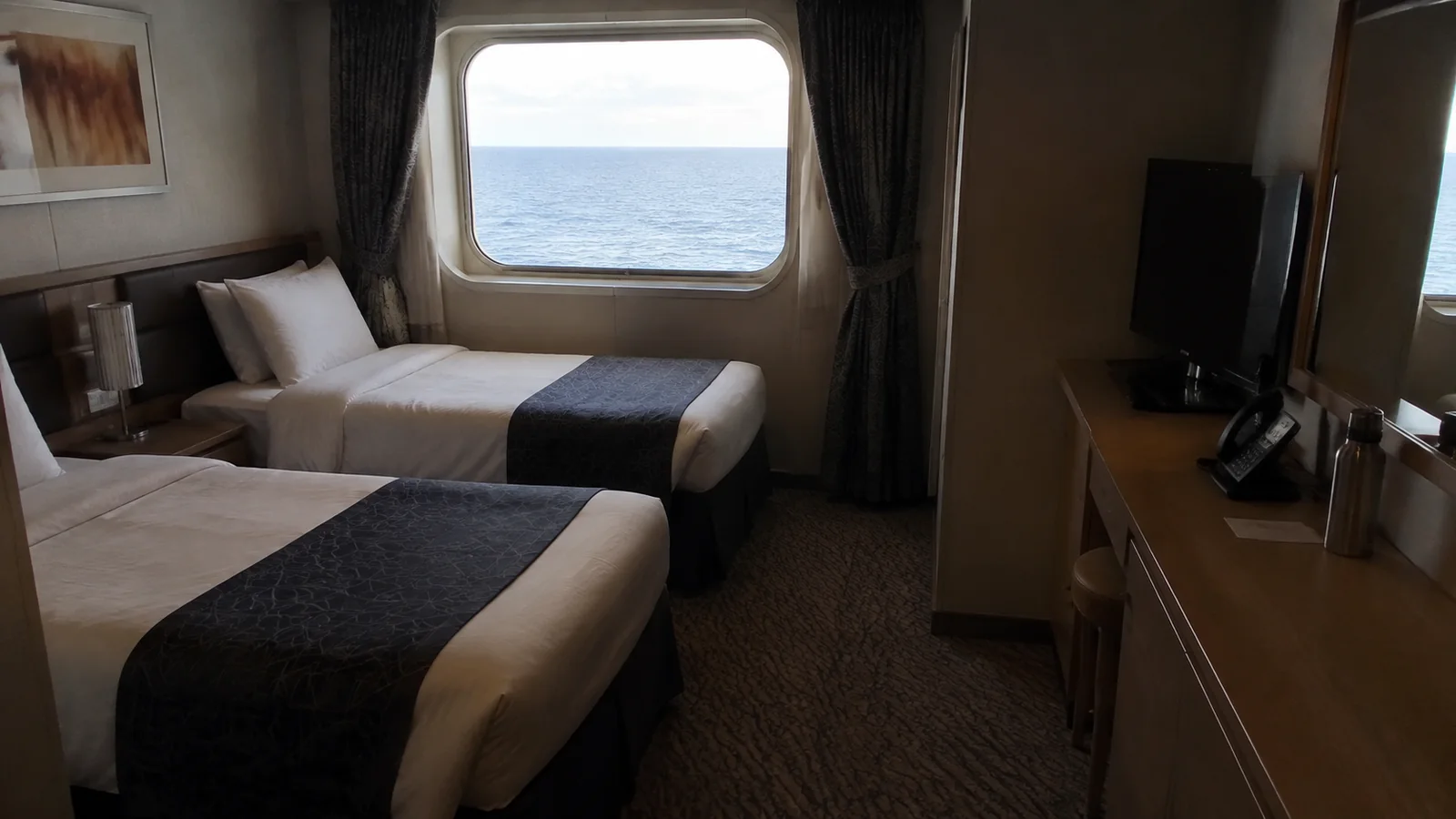 Twin beds and a wooden desk fill an oceanview stateroom.