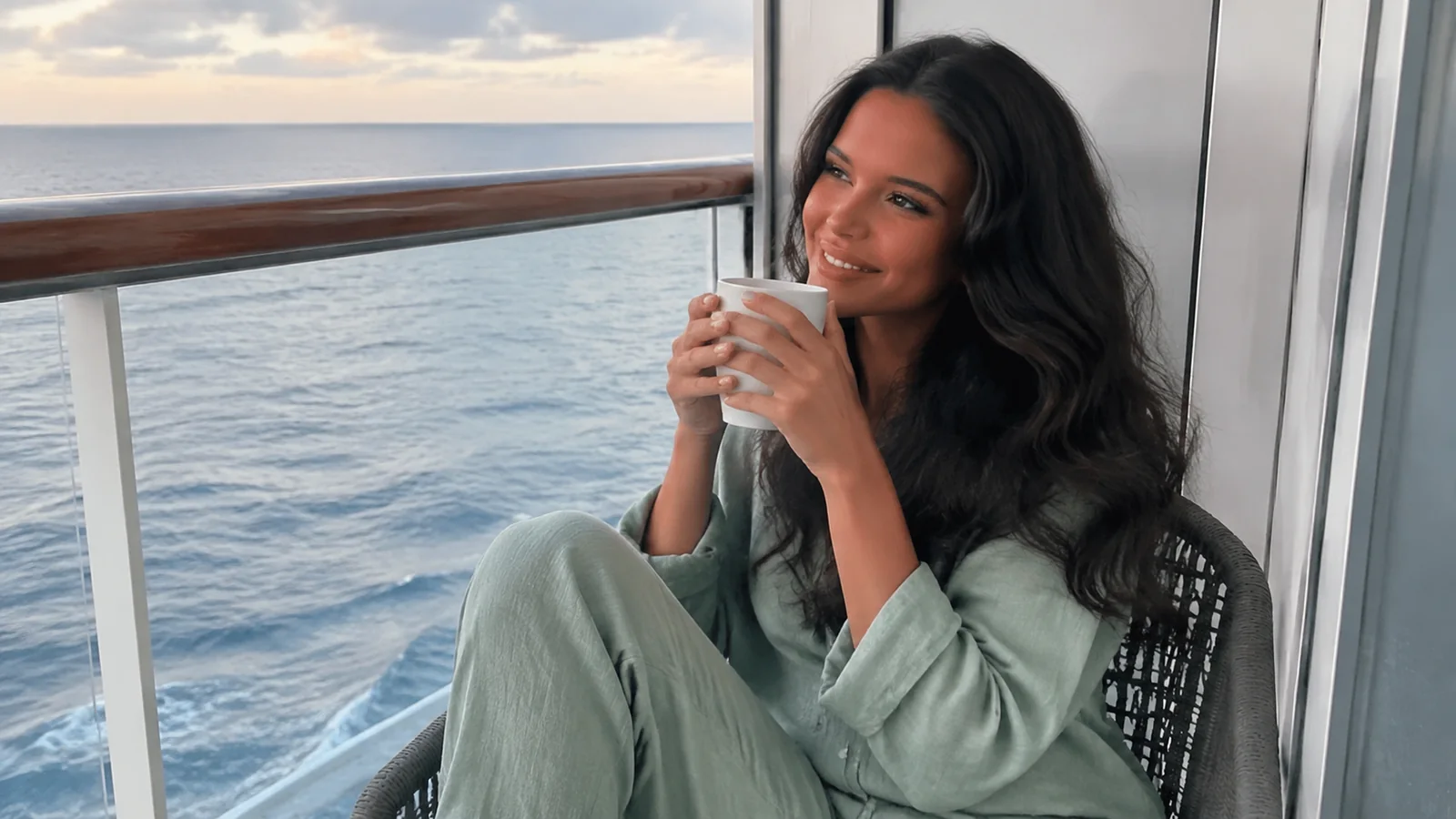 Woman holds a mug while seated on an ocean balcony.
