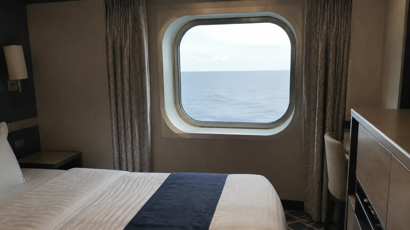 Stateroom window frames the sea above a bed and desk.