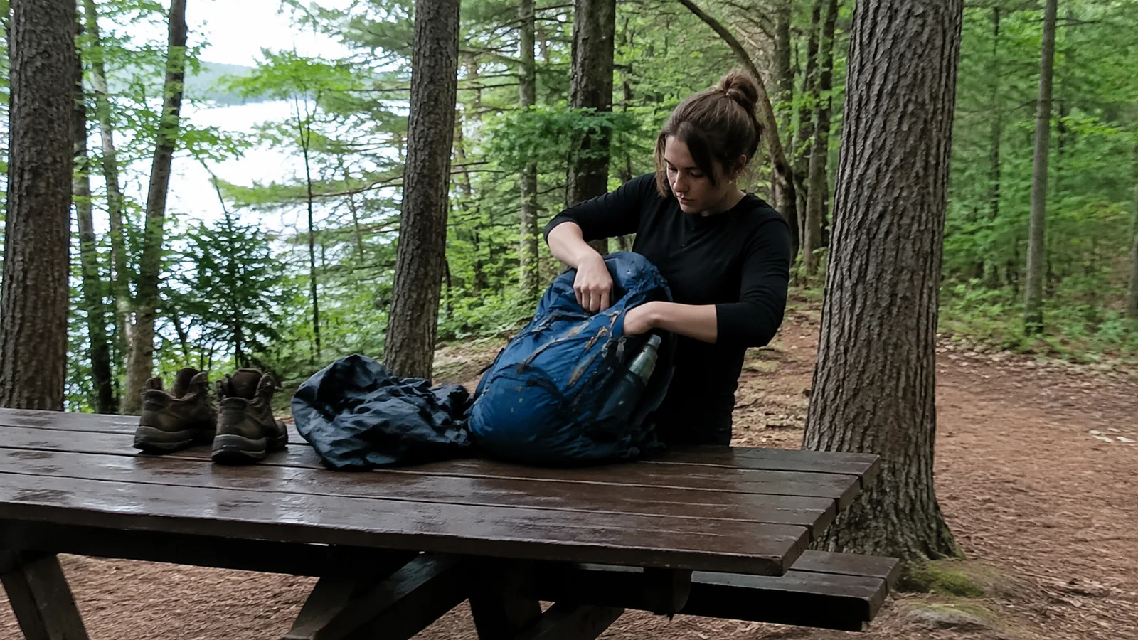 Camper packing a backpack at a forest picnic table beside hiking boots.