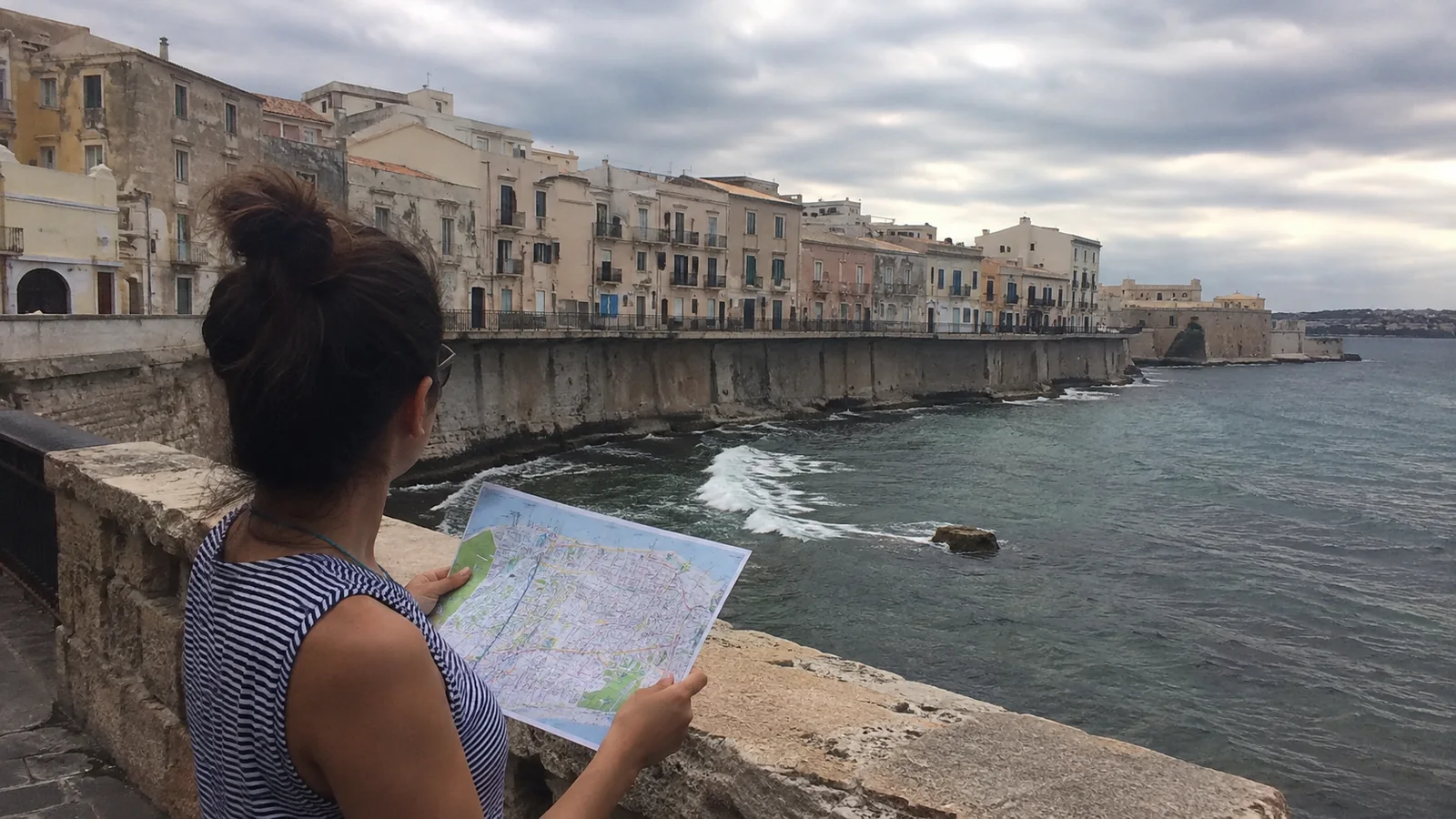 Woman holding a paper map at a waterfront overlooking a historic coastal city