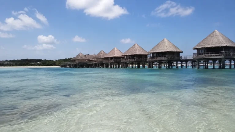 Thatched overwater bungalows stand above clear shallow turquoise water.