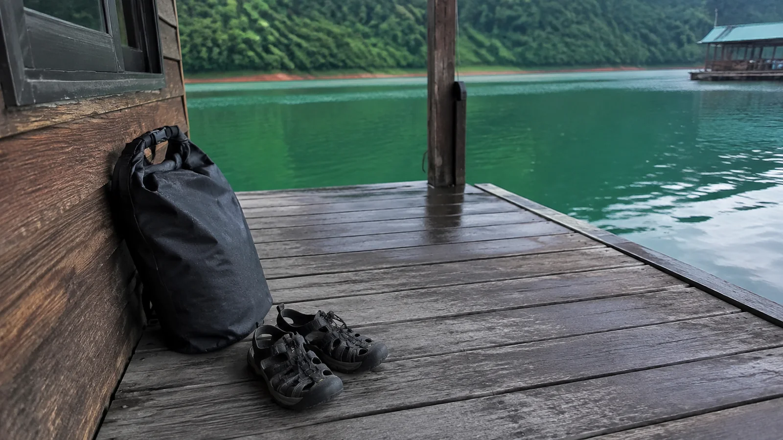 Dry bag and sandals on a wooden deck beside green water.