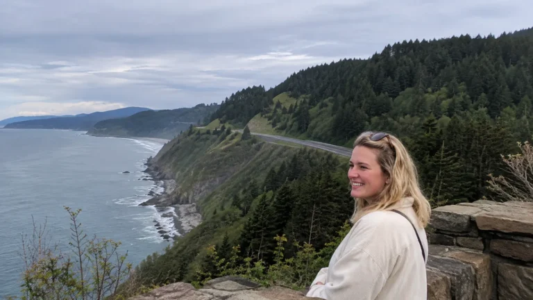 Woman at a coastal overlook above a winding road and forested cliffs.