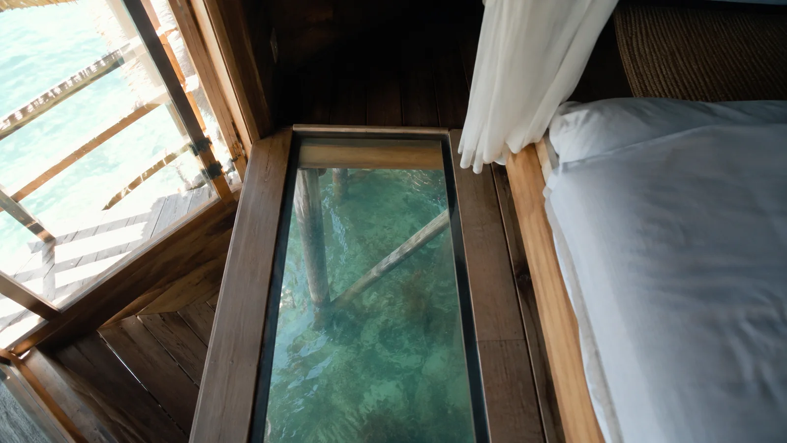 Glass floor panel beside a bed revealing water beneath an overwater bungalow.