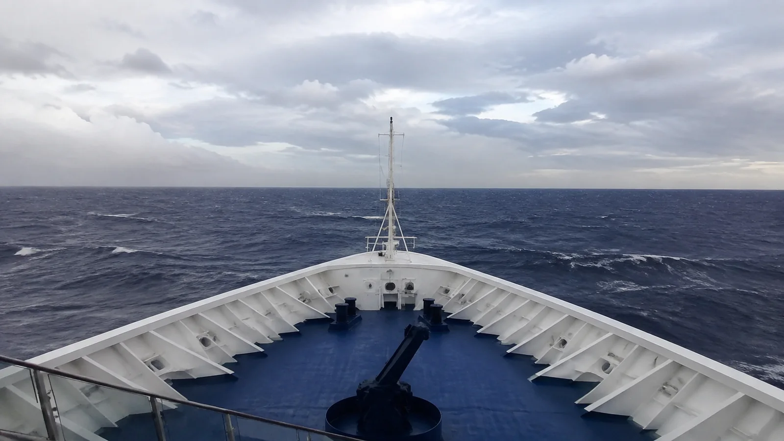 View over a ship's bow toward open ocean beneath gray clouds.