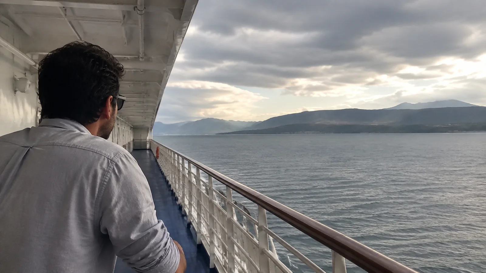 Passenger looking along a covered side deck toward a mountainous shore.