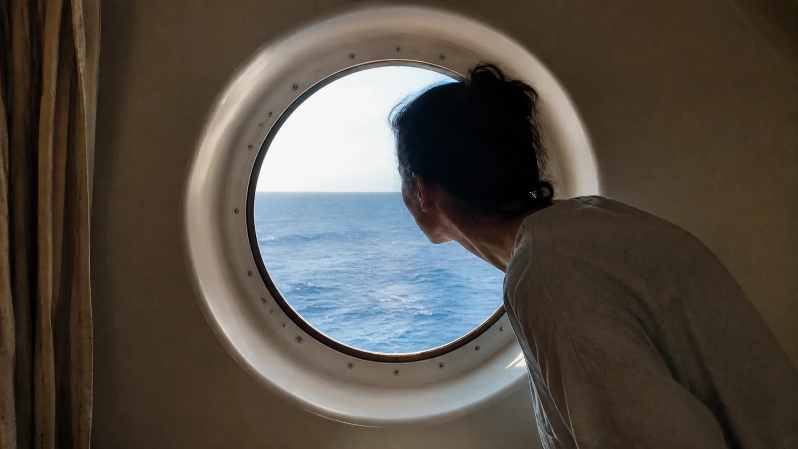 Passenger looking out to sea through a round cabin porthole.