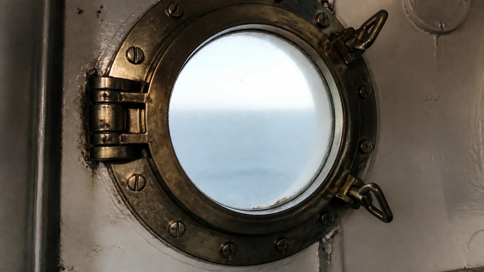 Brass-framed porthole with hinges and fastening dogs.